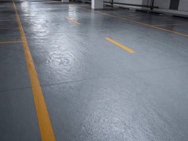 Wide overhead shot of an epoxy-coated parking level floor, safety-marking amber lane lines on mid-grey surface, existing overhead lighting, no vehicles or people, floor texture and joint expansion lines visible