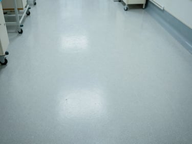 Overhead environmental shot of a hospital room floor, seamless light-grey epoxy surface, clinical ambient light, no people, hygienic finish visible