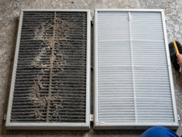 Overhead documentary shot of two AC filter panels side by side on a concrete floor under natural daylight — left panel clogged with thick dark dust, right panel freshly cleaned and pale white, technician's hand visible at edge holding a cleaning brush