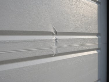Close-up of a bent garage door panel with visible crease damage, natural daylight, shot tight on the failure point