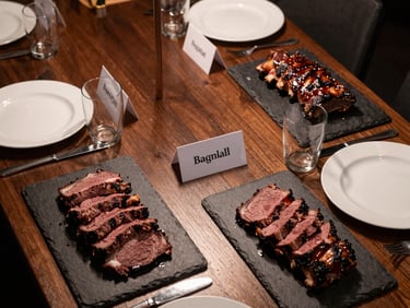 Overhead shot of a corporate dinner tablescape — slate boards with sliced smoked brisket and ribs, branded name cards, dark timber table, warm ambient light from above, no faces visible, clean and precise plating