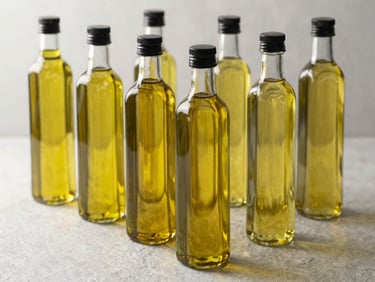 Clear glass bottles of golden vegetable and olive oils arranged neatly on a light slate background, soft natural light reflecting through the oil, clean and professional product shot