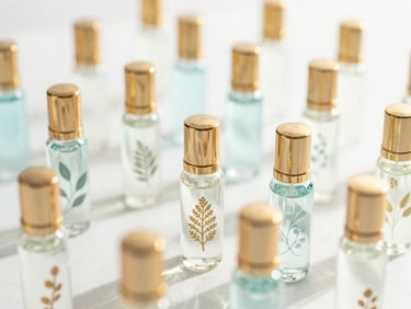 Delicate glass perfume vials with gold caps and botanical elements on a bright white surface, sun-drenched, high-key lighting, shallow depth of field.