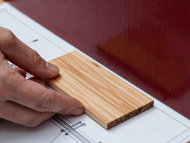 Macro close-up of hands matching a raw pine wood swatch against a deep crimson painted panel, architectural blueprint visible underneath, soft natural side light