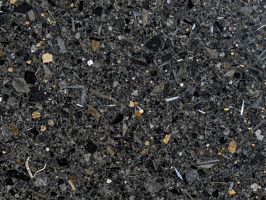 Extreme close-up overhead of a polished dark granite slab, dense interlocked mineral crystals in black, grey, and flecks of warm gold, north-facing daylight, no color correction, crystalline surface texture fully resolved