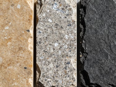 Extreme close-up overhead of three adjacent small stone samples — warm buff limestone, quartzite with silver-white shimmer, and dark charcoal slate with cleft texture — laid on a flat surface under north-facing daylight, no color correction, surface grain distinct across all three