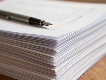 Close-up of a stack of neatly organized official documents, some open showing text, a fountain pen resting on top, soft natural daylight, warm cream tones, shallow depth of field.