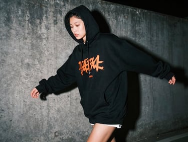 Model in oversized black streetwear hoodie with neon orange graphic print, urban concrete background, night flash photography