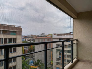 Wide apartment balcony in daylight with a fine-mesh protective net installed along the open railing, city buildings softly visible through the net in the background
