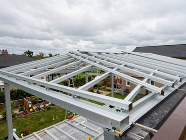 Wide shot of a terrace roof structure mid-installation, aluminum rafters spanning across, crew member securing a purlin from a ladder, overcast exterior daylight, residential garden below