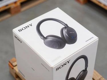 Close-up overhead view of sealed Sony WH-1000XM and Samsung Galaxy Buds retail boxes stacked on a pallet shelf, serial number stickers visible on box edges, clean warehouse daylight from above, plain concrete floor in background