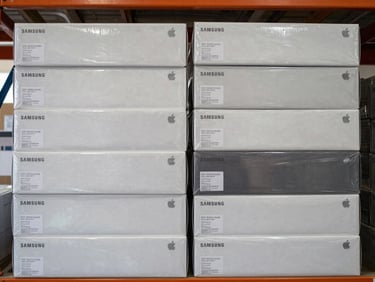 Overhead close-up of neatly stacked sealed Samsung and Apple case boxes arranged in two rows on a warehouse shelf, printed authentication seals visible, clean daylight from the left, neutral background