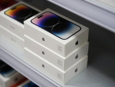 Overhead close-up of sealed Apple iPhone boxes stacked three high on a white warehouse shelf, manufacturer authentication seal visible on each box, studio strobe lighting on product