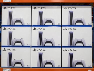 Wide overhead shot of sealed laptop boxes and PS5 console boxes stacked side by side on a distribution center shelf, clean daylight, authentication seal labels visible