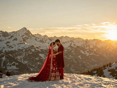An Indian couple in flowing crimson couture on a snow-capped Swiss mountain peak, dramatic golden hour sun, wide atmospheric landscape.