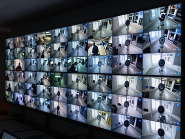 Command center monitor wall displaying a grid of live CCTV feeds from multiple camera angles, low ambient light, blue status indicators active, no identifiable individuals on screens