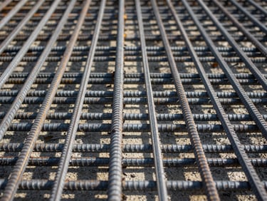 Macro flat-lay of interlocking steel reinforcement bars laid out in a precise grid pattern for concrete foundation, harsh midday sun casting sharp geometric shadows on the ground.