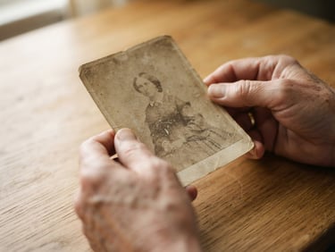 Macro close-up of hands holding a fragile, faded 19th-century black-and-white photograph over a clean wooden table, soft warm window light casting gentle directional shadows, showcasing rich paper texture.