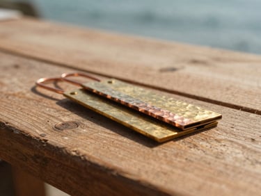Close-up of hand-hammered copper wire and polished brass sheets resting on a rustic wooden workbench, warm direct sunlight casting sharp shadows.