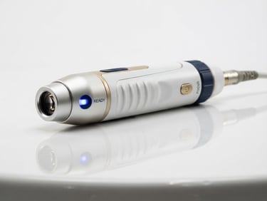 Macro shot of a sleek medical laser handpiece resting on a polished white clinical surface, soft gold and navy blue reflections, high-tech glowing indicator light, premium product photography.