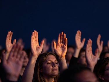 Close-up of hands raised in worship during an evening service, subtle fiery-orange light leaks, deep navy background.
