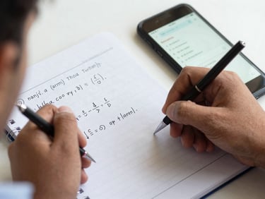 Close-up of a Pakistani student's hand solving a physics equation in a notebook next to a mobile screen showing test results, bright natural light