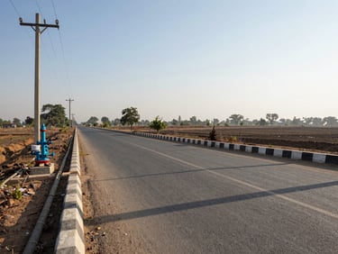 Panoramic environmental shot of township infrastructure at Jhajjar — wide internal road with visible underground utility conduits alongside the kerb, installed power poles and water supply headers visible, early morning natural daylight casting long parallel shadows across the road surface, no figures, low horizon showing open land beyond