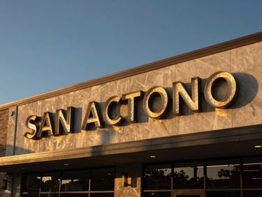 Wide environmental shot of illuminated channel letters mounted on a commercial building facade at golden hour, letters casting warm glow against dark sky, San Antonio streetscape visible below