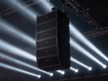 Concert-grade line array sound system suspended from a truss, dramatic stage lighting beams cutting through haze, low-key background