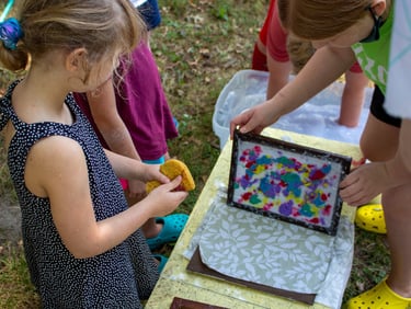 Group of young children holding a colorful screen above a piece of fabric while making paper.