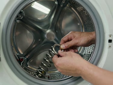 Extreme close-up of a technician's hands replacing a broken door spring on a washing machine drum, fluorescent garage light, worn concrete floor visible