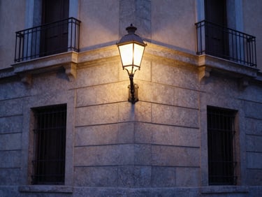 Quiet neighborhood corner in Madrid at dusk, elegant street lamp lighting up a stone facade, soft deep indigo and warm sand-beige tones