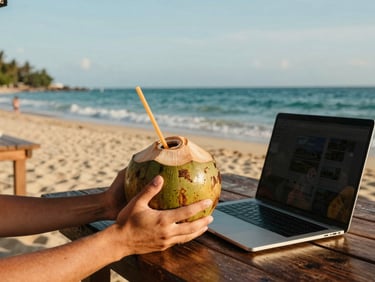 Hands holding a cold coconut at a beachside kiosk, laptop open on a wooden table, ocean background.