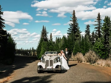 Bride and groom posing beside a vintage car on a scenic forest road, captured by Fred Art Studio.