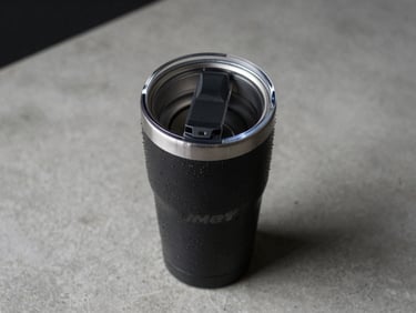 Close-up overhead studio shot of a Yeti Rambler tumbler on a raw concrete desk surface, cold studio lighting, water condensation visible on the stainless steel, no hands, no people, black and silver tones dominant