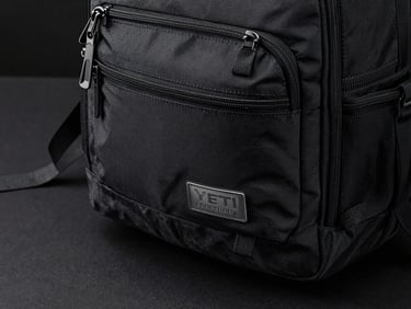 Close-up of a Yeti Crossroads backpack on a matte black desk surface, side pocket zipper and webbing strap in sharp focus, studio lighting from above, no lifestyle context