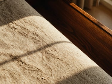 Close-up of tactile materials, warm sand-toned linen and polished dark wood in a sunlit interior, soft shadows, editorial framing.