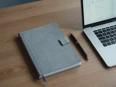 Overhead shot of a minimalist wooden desk with a neat stack of grey notebooks, a black pen, and a laptop showing clean spreadsheets, cool diffused daylight, quiet editorial framing.
