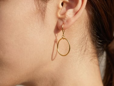 Macro photography of minimalist gold-plated steel earrings on a woman's ear, soft shadows, warm editorial aesthetic.