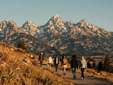A small group of friends walking on a sun-drenched mountain trail with Teton peaks rising in the background.