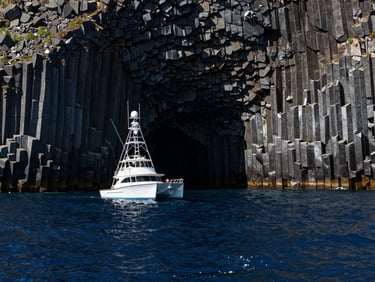 Wide environmental shot of the Mitsio basaltic cliffs rising from a deep navy blue ocean, high contrast midday sun reflecting off the water, a professional sport fishing catamaran positioned off-center left