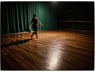 Wide panoramic view of an elegant, dimly lit dance studio with polished dark wood floors, warm tungsten lighting reflecting off the surface, soft emerald drapes in the background, negative space on the right.