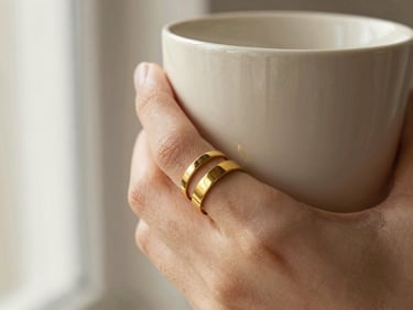 Stacked gold band rings on fingers holding a cream ceramic cup, soft shadows