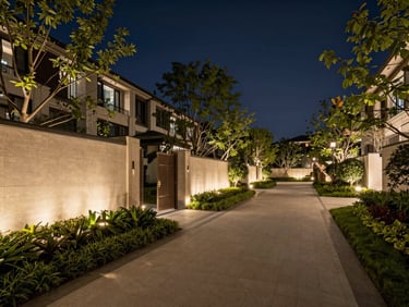 Wide environmental shot of a luxury gated residential perimeter at dusk — illuminated boundary wall with mature landscaping, a long paved walkway receding into the distance, warm uplighting on trees, deep navy sky, no people