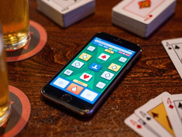 Overhead shot of a mobile phone displaying a smooth, active card game, surrounded by beer coasters and card decks on a rich wooden table, vibrant neon glow, shallow depth of field