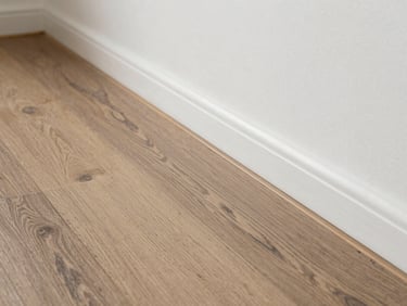 Close-up, bright natural light photograph of freshly laid luxury vinyl plank flooring meeting a perfectly painted white baseboard transition.