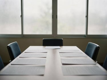 Empty quiet meeting room with two chairs facing each other across a narrow table, documents spread flat, natural daylight through frosted glass partition, no people visible
