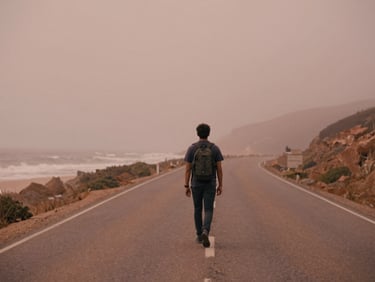 Cinematic 16:9 video still of a lone traveler on a misty coastal highway, muted terracotta tones, shallow depth of field, film grain.