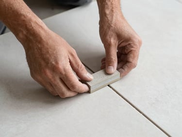 Overhead close-up of a tile installer placing a spacer between two large-format porcelain tiles, crisp gray grout lines, natural side lighting.