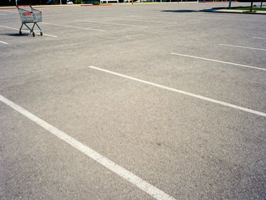 Empty parking lot at midday in hard summer sun, early 2000s, digital grain, a lone shopping cart at the far edge, flat harsh shadows